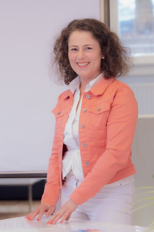 jenny-spiller-happy-mom-coaching frau mit rosaner jacke zeigt mit beiden Händen eine Grafik auf dem Tisch